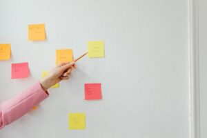A hand pointing with a pencil at a yellow sticky note on a wall covered with colorful sticky notes, each containing handwritten tasks and ideas