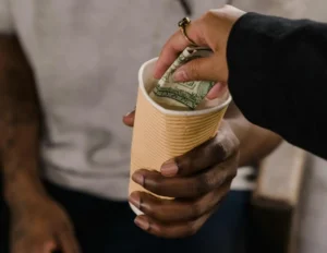 A close-up of a hand placing a folded dollar bill into a paper cup held by another person